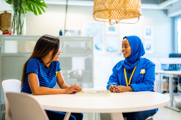 Zwei IKEA Mitarbeitende unterhalten sich an einem modernen runden Tisch in einem hellen, zeitgemäßen Arbeitsbereich, umgeben von Pflanzen und Tageslicht.