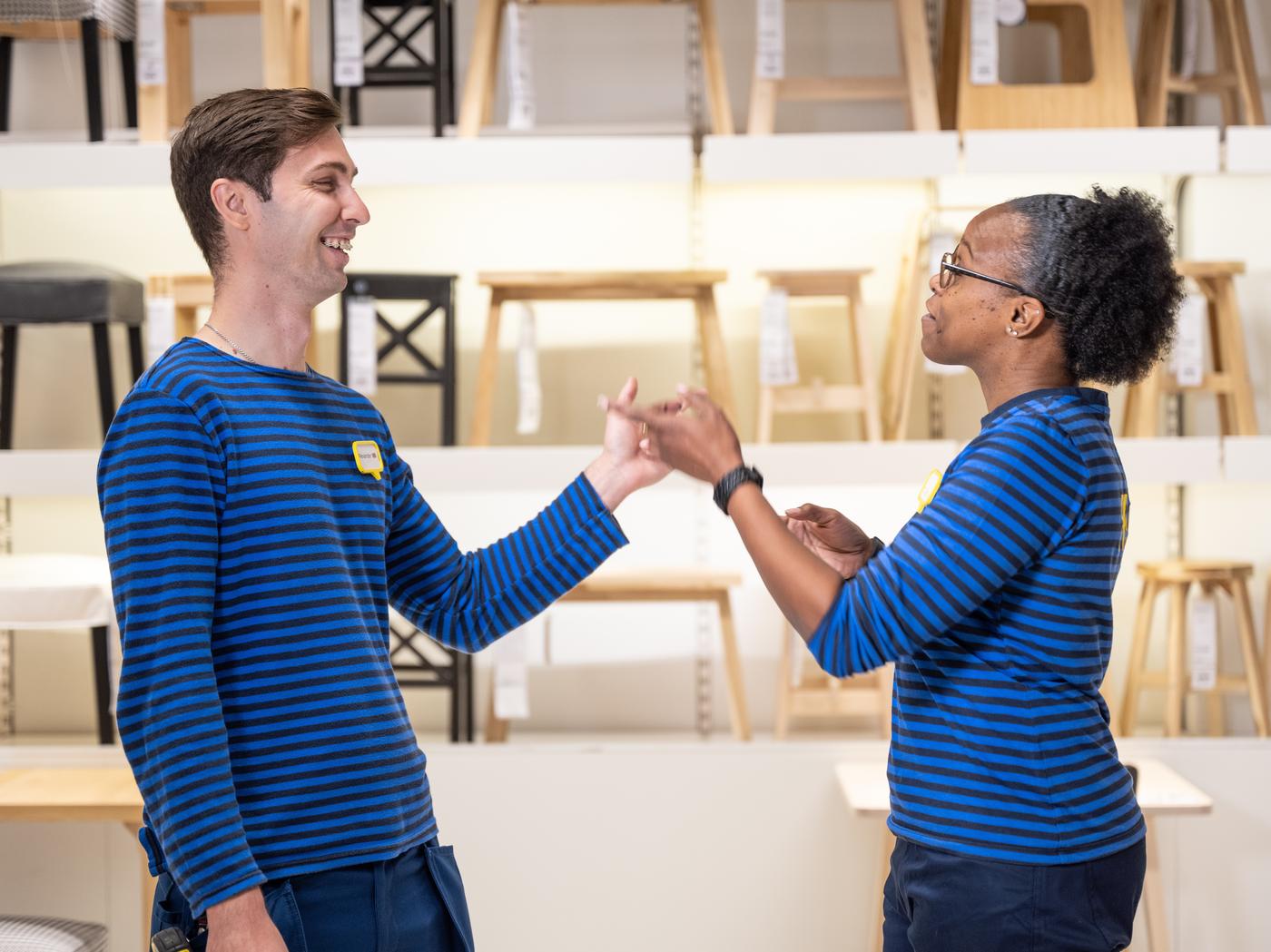 Zwei IKEA Mitarbeiter:innen in blaue gestreiften Oberteilen unterhalten sich vor der Möbelausstellung im Einrichtugshaus. Im Hintergrund sind Hocker zu erkennen.