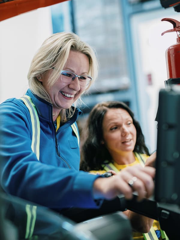 Zwei IKEA Mitarbeiterinnen lächeln bei einer Schulung in einem Lagerbereich, eine zeigt etwas an einem Gerät.