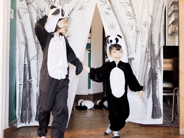 Zwei Kinder in einem Pandabärkostüm an einem Theaterabend verbeugen sich auf einer Bühne, die mithilfe eines geschmückten BJÄRSEN Duschvorhangs gestaltet wurde.
