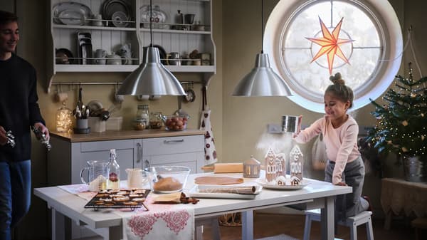 Zwei Kinder setzen ein Pfefferkuchenhaus zusammen und schmücken es mit Puderzucker, der darüber wie Schnee verteilt wird.
