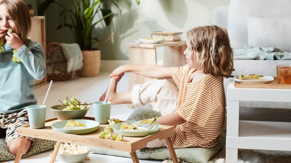 Zwei Kinder sitzen im Wohnzimmer auf dem Boden. Vor ihnen steht ein Tablett aus Bambus mit Obstschnitzen und Getränken.