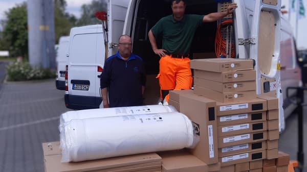 Zwei Männer stehen vor einem offenen Lieferwagen, umgeben von gestapelten IKEA-Kartons und gerollten Matratzen. Einer trägt eine dunkelblaue Jacke, der andere ein grünes Polohemd mit leuchtend orangefarbener Arbeitshose. Im Hintergrund sind weitere weisse Transporter zu sehen.  