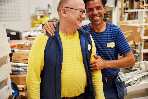 Zwei männliche IKEA Mitarbeiter, die sich lachend im Arm halten. Im Hintergrund befinden sich sämtliche Utensilien, sowie Kartons.