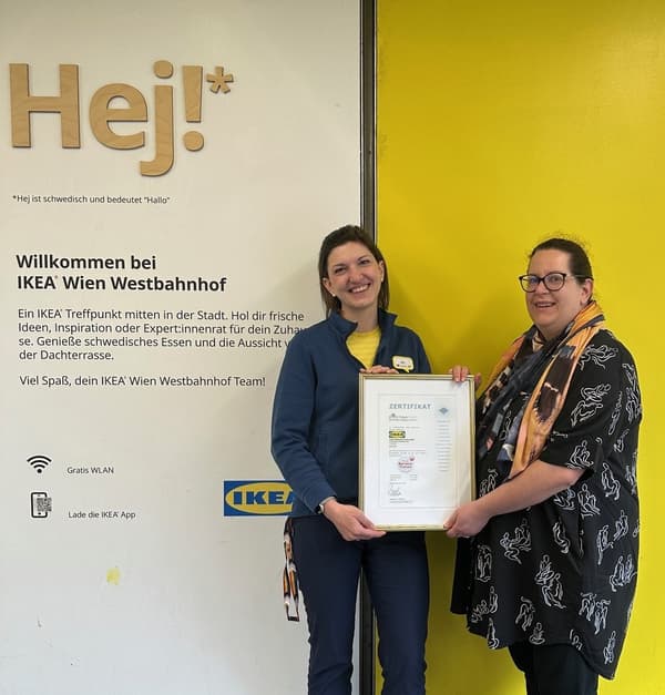 Zwei Mitarbeiterinnen des IKEA Wien Westbahnhof stehen lächelnd vor einer Begrüßungswand und halten gemeinsam ein Zertifikat – Zeichen für Auszeichnung oder Anerkennung.
