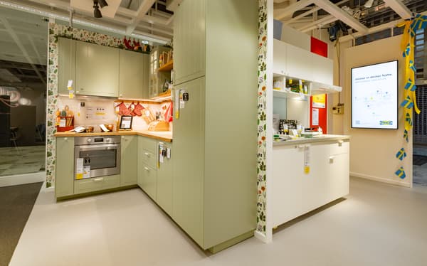 Zwei Musterküchen bei IKEA. Links eine U-Küche in hellem Grün mit floraler Tapete, rechts eine moderne weiße Küche mit offenen Regalen. Auf einem Monitor wird IKEA Küchenplanung beworben.