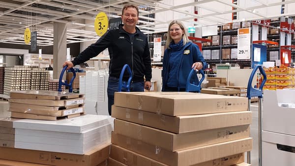 Zwei Personen befinden sich in einem IKEA Möbelhaus, in der Selbedienungshalle und haben jeweils einen Transportwagen mit mehreren flach verpackten Möbelkartons vor sich stehen. Die Umgebung ist hell und geräumig, mit hohen Regalen, Preisschildern und Lagerhaus-Atmosphäre.