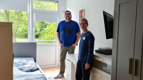 Zwei Personen befinden sich in einem kleinen Zimmer in der Regelwohngruppe des DRK. Das Zimmer ist mit einem Bett, einem Schrank, einer Kommode und einem Schreibtisch eingerichtet. 