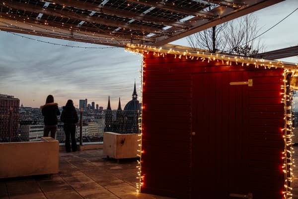 Zwei Personen schauen von einem Dach aus auf die Skyline der Stadt, im Hintergund ist ein Weihnachlich gemschmückter Stand zu sehen.