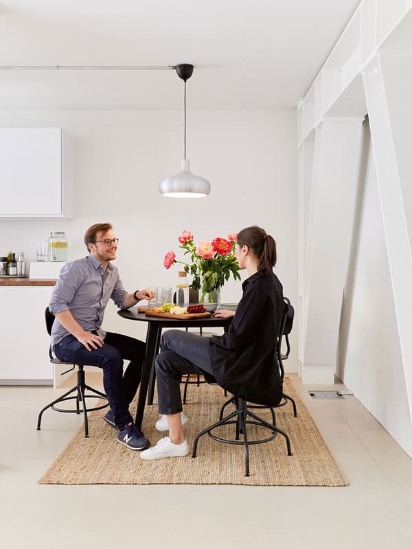 Zwei Personen sitzen lachend an einem runden Esstisch in einer Kaffeeküche einer Anwaltskanzlei. Auf dem Tisch stehen Snacks und ein Blumenstrauss in einer Vase.