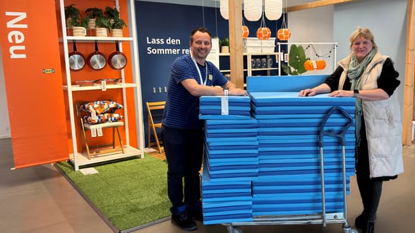 Zwei Personen stehen im Eingangsbereich vom IKEA Wuppertal  zusammen. Einer trägt IKEA-Dienstkleidung. Eine Frau vom Kinderschutzbund nimmt die Spielmatten entgegen. Alle blicken in die Kamera und lächeln.