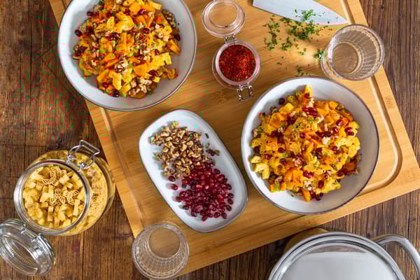 Zwei Portionen herbstliche Kürbis-Pasta mit Walnüssen stehen angerichtet mit Granatapfelkernen auf einem Holztisch.