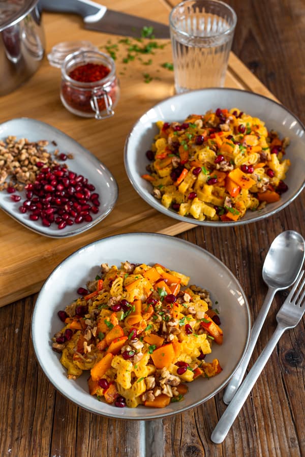 Zwei Portionen herbstliche Kürbis-Pasta mit Walnüssen stehen angerichtet mit Granatapfelkernen auf einem Holztisch.