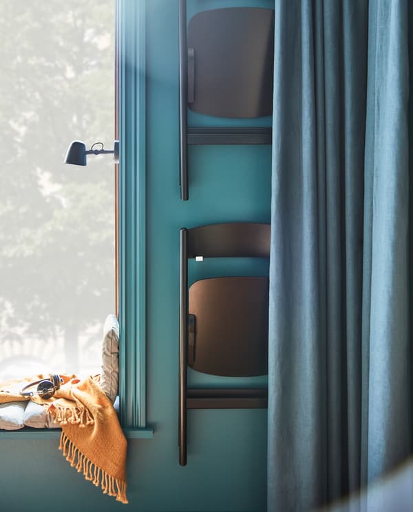 Zwei schwarze Klappstühle hängen an Haken an der Wand neben einem Fenster. Sie lassen sich hinter blauen Gardinen leicht verbergen.