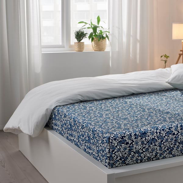 Bedroom with white bedding; blue floral-printed flat sheet; white pillows; two potted plants on windowsill.