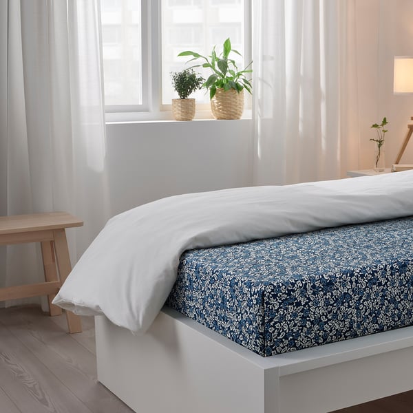 Bedroom scene with a white bed. The bed has a patterned blue sheet and a white pillowcase. Two potted plants are on the windowsill behind the bed.