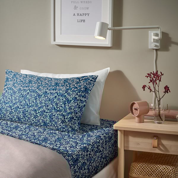 Cosy bedroom with blue floral duvet, white pillow, pink clock, flowers, books, motivational poster, and wall lamp.