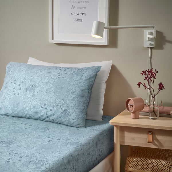 Cosy bed with blue floral print, pink clock, flowers, and wall light. Or, even shorter: blue floral bed with pink clock and flowers.