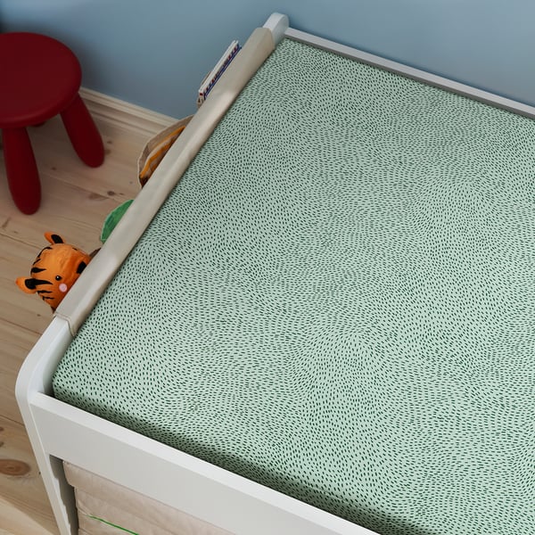 A crib with a green, dot-patterned fitted sheet. A stuffed tiger and a red stool are nearby.