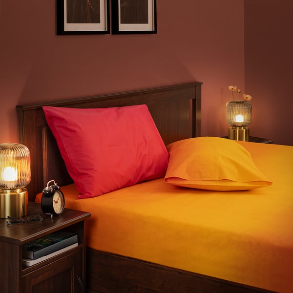 Bedroom with wooden headboard, orange sheets, pink pillows, lamps, clock. Vibrant, fresh atmosphere.