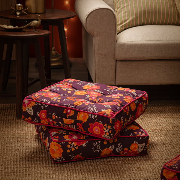 Bright floral-patterned purple floor cushion, with a handle, sits beside a beige sofa, adding extra seating.