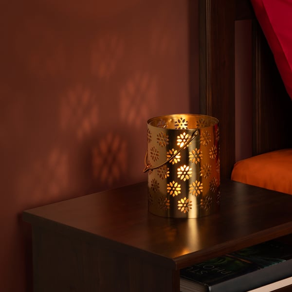Gold decorative lantern on a table, casting warm light and intricate floral shadows. Part of the AROMATISK collection.