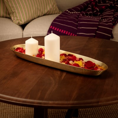 Gold-colored oval dish with candles and rose petals sits on a wooden table, bordered by a cushioned bench with striped pillows.