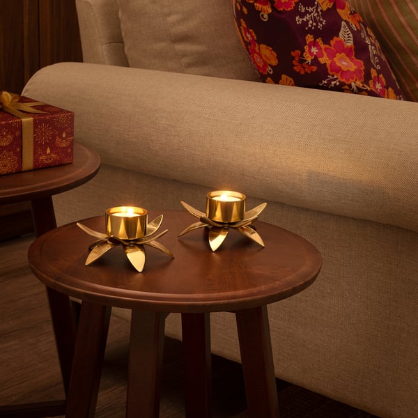 Two lit golden lotus tealight holders on a wooden table beside a beige couch with a floral pillow.