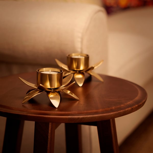 Two AROMATISK gold tealight holders on wooden table, one lit, one unlit, small and elegant, reflecting flame, intricate lotus shape.