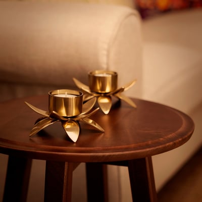 Two AROMATISK gold tealight holders on wooden table, one lit, one unlit, small and elegant, reflecting flame, intricate lotus shape.