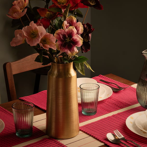 A table set with red placemats, white plates, clear glasses, forks, and a gold vase holding colourful flowers.