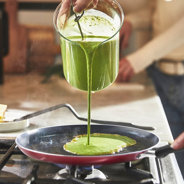 Person pouring green batter from a glass jug into a red non-stick frying pan on a gas stove.