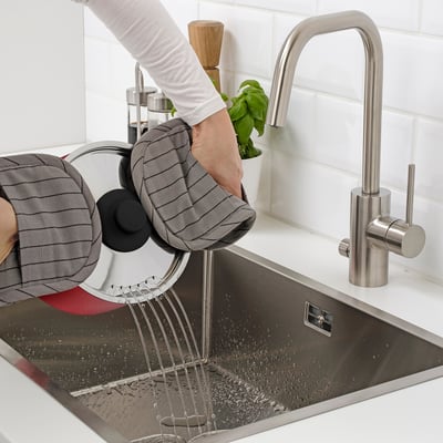 A hand holding a grey pot with a strainer inside is washing it in a stainless steel sink with running water from a modern tap.