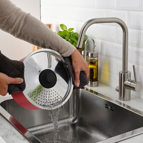 Person washing BAKÅTVOLT pot in sink, water pouring through lid holes.