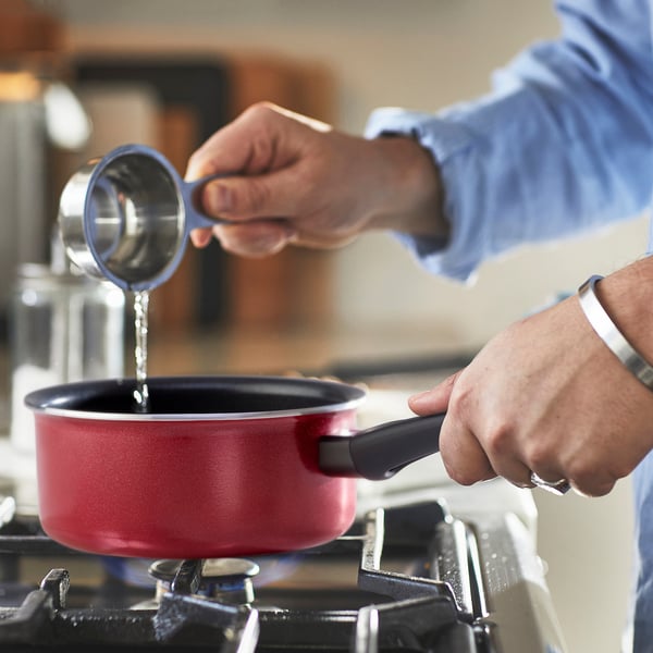 Person pours liquid into red saucepan on stove, using measurement spoon. Saucepan has a black handle and is placed on a gas burner.