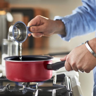 Person pours liquid into red saucepan on stove, using measurement spoon. Saucepan has a black handle and is placed on a gas burner.