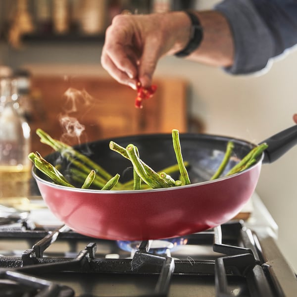A hand in a glove adds chilli to a red frying pan with green beans on a gas stove.