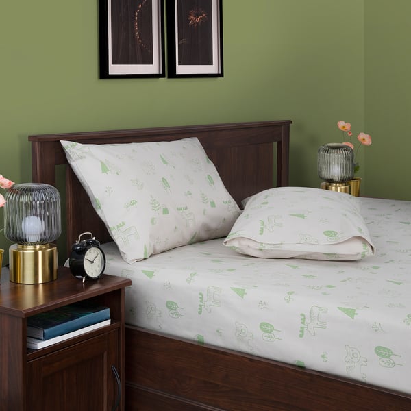 Green bedding with nature-themed print on a wooden bed. Includes pillows, bedside lamp, alarm clock, and vase with flowers on a bedside table.