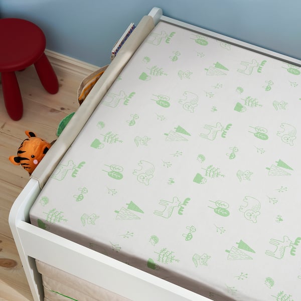 A white baby cot with a green patterned mattress featuring forest animals, set in a calm blue room with a red stool nearby.