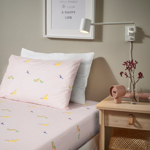 A pink bed with bardröm bedding featuring flying unicorns. The bedding is made of cotton and lyocell. A side table with a lamp and vase is next to it.