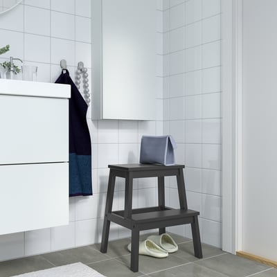 Modern bathroom with BEKVÄM black wood step stool, blue towel, white slippers. Tiled walls and floor.