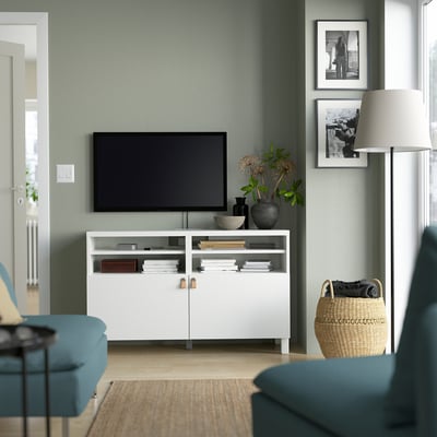 Modern living room featuring BESTÅ tv bench, flat-screen tv, blue sofa, plants, and framed photos against sage-green walls.