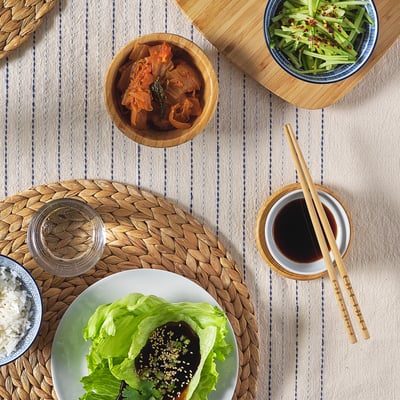 A meal setup with wooden bowls, chopsticks, and plates; includes lettuce wraps, rice, kimchi, and cucumber salad on a striped tablecloth.