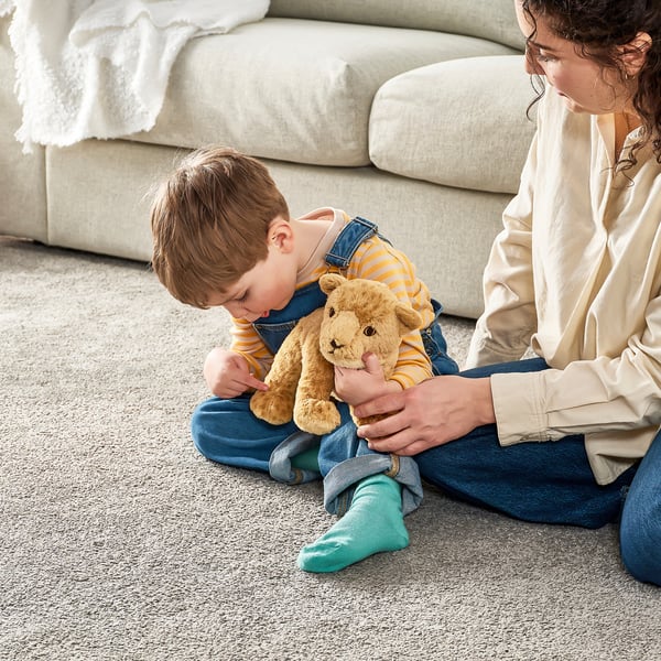 A child sits with a person, hugging a soft lion cub toy. The toy is designed for comfort and play, suitable for ages 0+.