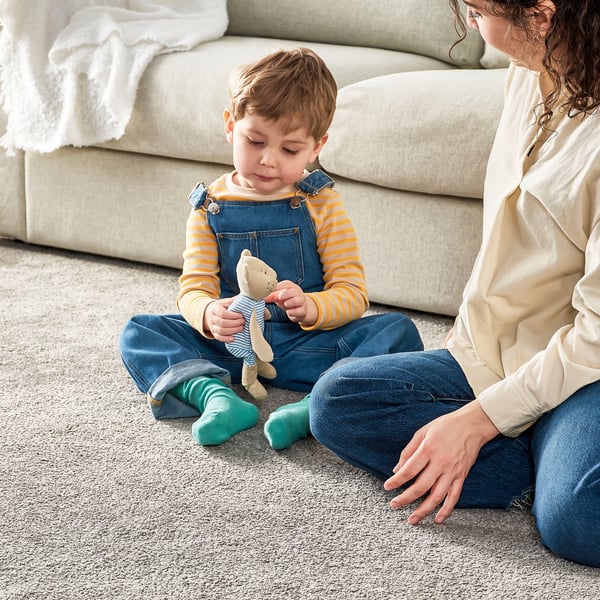 Young boy in overalls sits on floor, holding small teddy bear, as person in jeans sits beside the person.