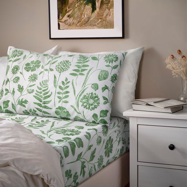 Bed with green FÄRGARE floral sheet set on white bedding, including pillowcases, on bedside table with decor.