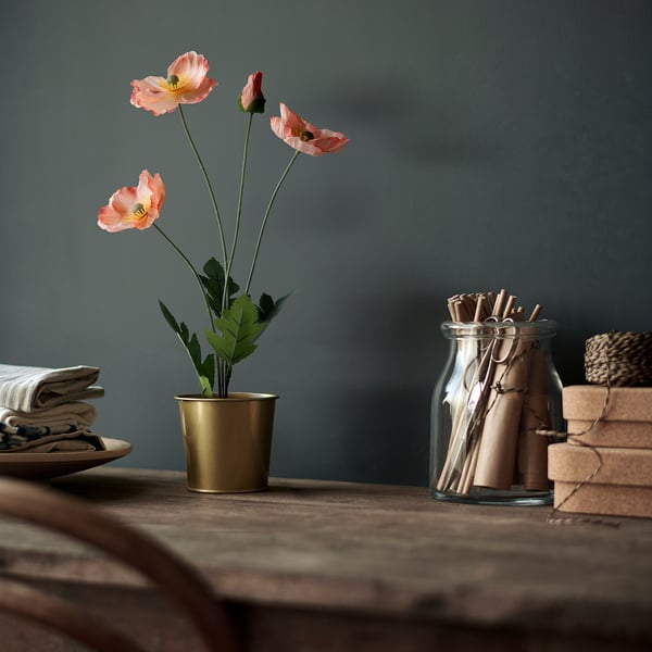 A gold pot with pink artificial poppies and green leaves sits on a wooden table. Beside it, a glass jar holds rolled paper.