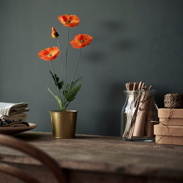 Vibrant orange artificial poppies in a gold pot on a wooden table, accompanied by folded napkins, cinnamon sticks, and books.