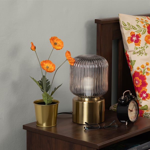 Bedside display: golden pot with bright orange poppies, cylindrical ribbed lamp, classic black alarm clock, colourful floral pillow on wooden drawer.