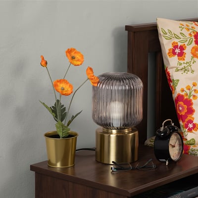 Bedside display: golden pot with bright orange poppies, cylindrical ribbed lamp, classic black alarm clock, colourful floral pillow on wooden drawer.